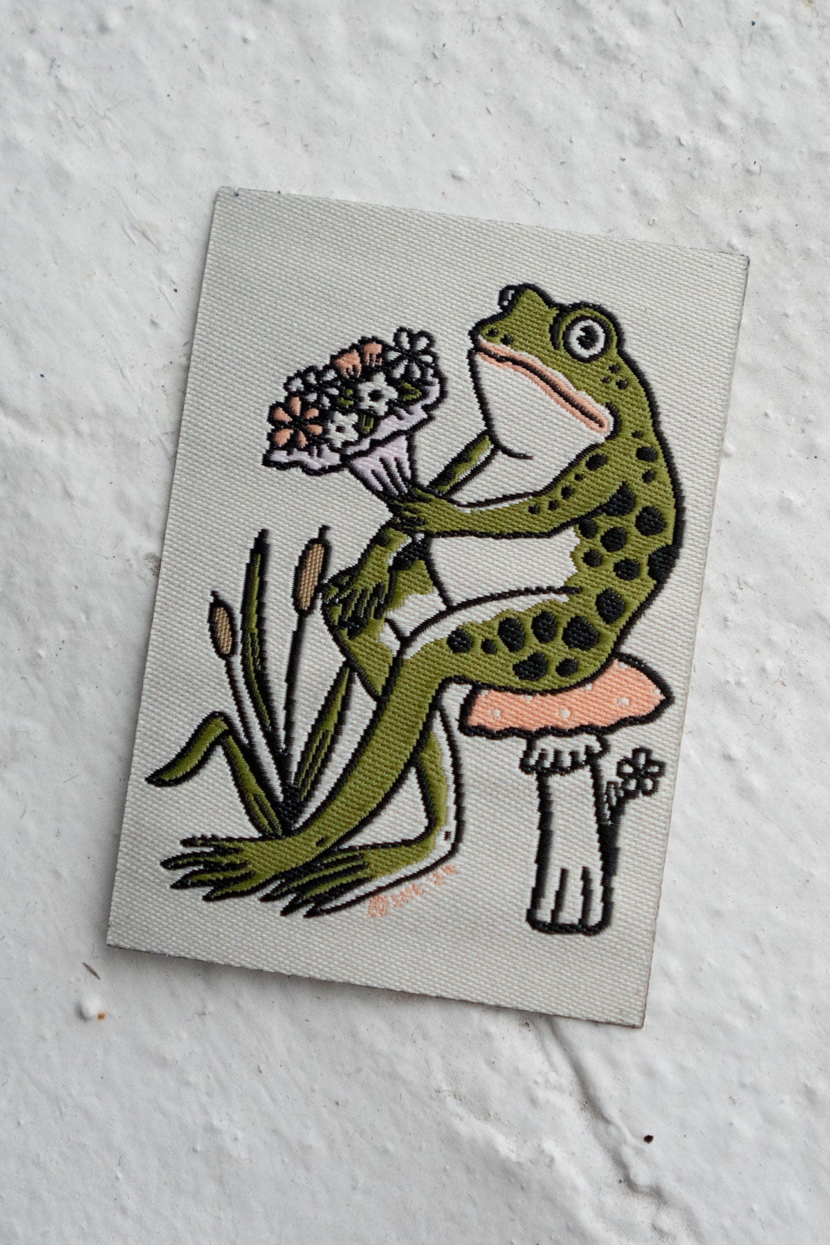 Woven patch with an image of a frog sitting on a mushroom and holding a bouquet of flowers