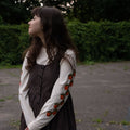 Woman in a brown dress layerd over an off-white long sleeve t-shirt featuring fruit patterns standing outdoors.