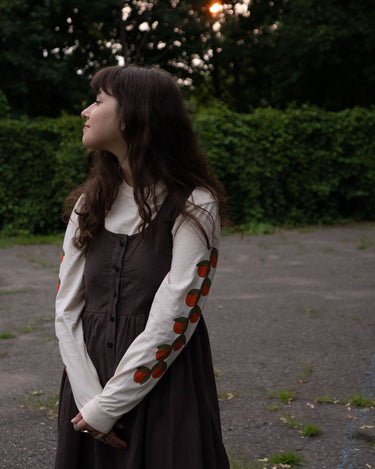 Woman in a brown dress layerd over an off-white long sleeve t-shirt featuring fruit patterns standing outdoors.