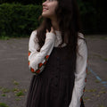 Woman in a brown dress and off-white long sleeved shirt with orange patterns standing outdoors.