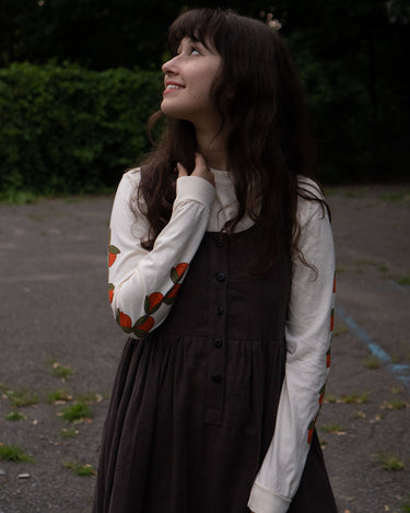 Woman in a brown dress and off-white long sleeved shirt with orange patterns standing outdoors.