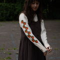 Woman wearing an off-white long-sleeve shirt with orange designs, standing outdoors.