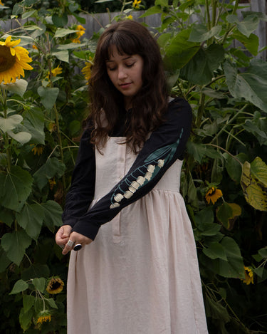 Woman in a garden with sunflowers wearing a dress and blacklong-sleeved t-shirt with floral print sleeves.