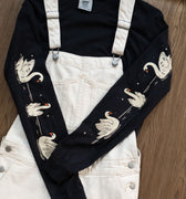 Black long-sleeved t-shirt with swans printed down each arm layered under off-white overalls.