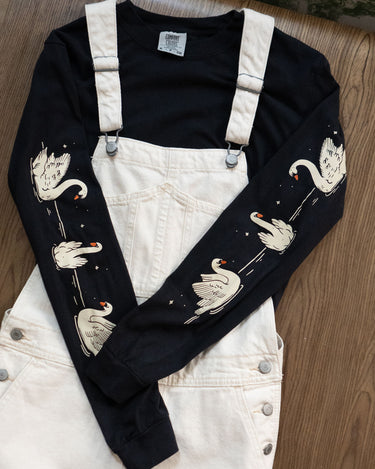 Black long-sleeved t-shirt with swans printed down each arm layered under off-white overalls.