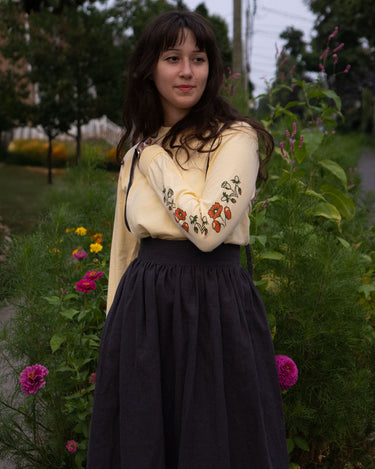 Woman wearing a long-sleeved shirt with floral print sleeves and dark skirt standing in a garden.