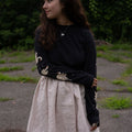 Woman wearing a black long-sleeved t-shirt with swans printed on the sleeves and beige skirt standing outdoors with greenery in the background
