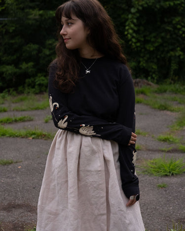 Woman wearing a black long-sleeved t-shirt with swans printed on the sleeves and beige skirt standing outdoors with greenery in the background
