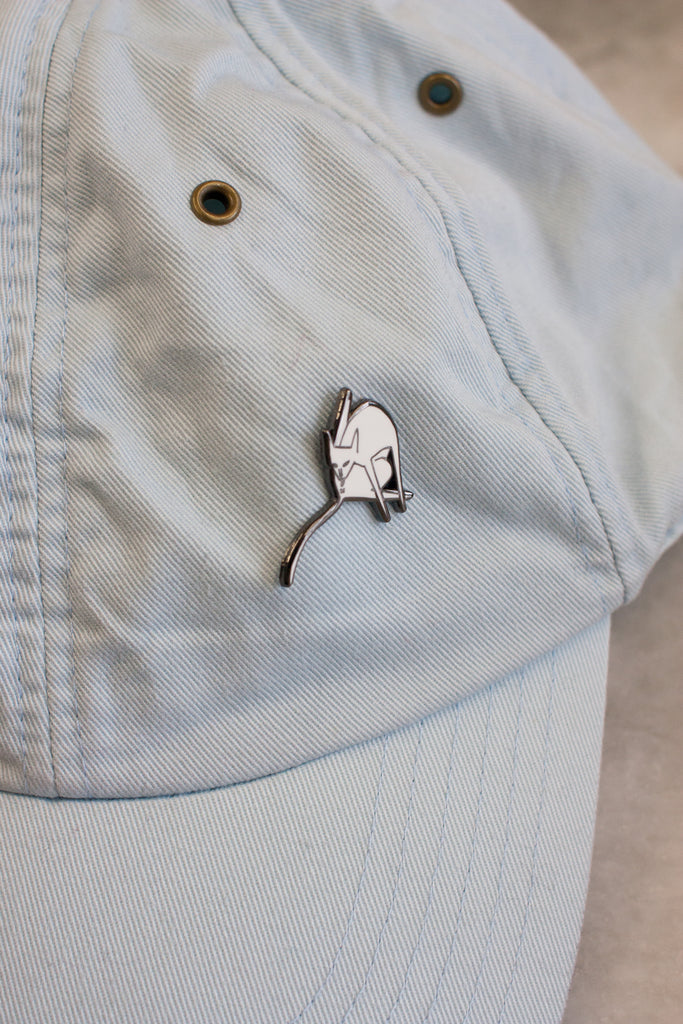 lapel pin of white cat licking itself with leg up and a long tail pinned on a pale blue baseball cap
