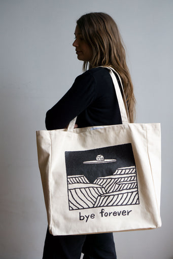 person holding natural canvas tote bag with square image in black of house floating on island with text reading "Bye forever" below illustration