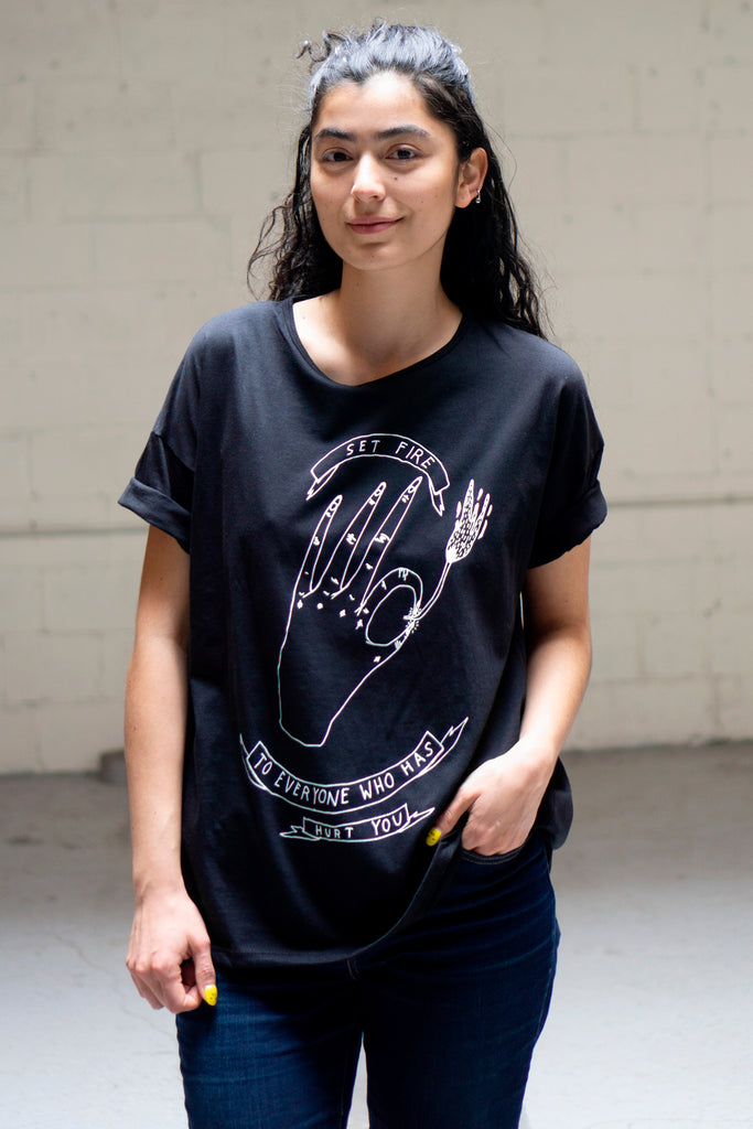 Model wearing loose fitting black t-shirt with illustrated print of a hand lighting a match and readin "set fire to everyone who has hurt you"