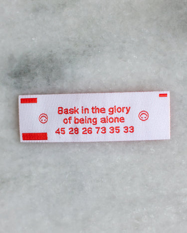 Small rectangular woven patch in the style of a fortune cookie insert, with red text reading "bask in the glory of being alone"