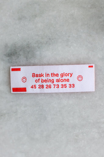 Small rectangular woven patch in the style of a fortune cookie insert, with red text reading "bask in the glory of being alone"