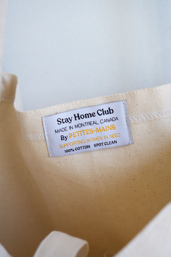 detail of woven tag inside tote bag reading "Stay home club Made in Montreal, Canada by Petites-Mains Supporting Women in Need 100% cotton spot clean"