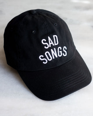 black baseball hat with white embroidered text reading SAD SONGS