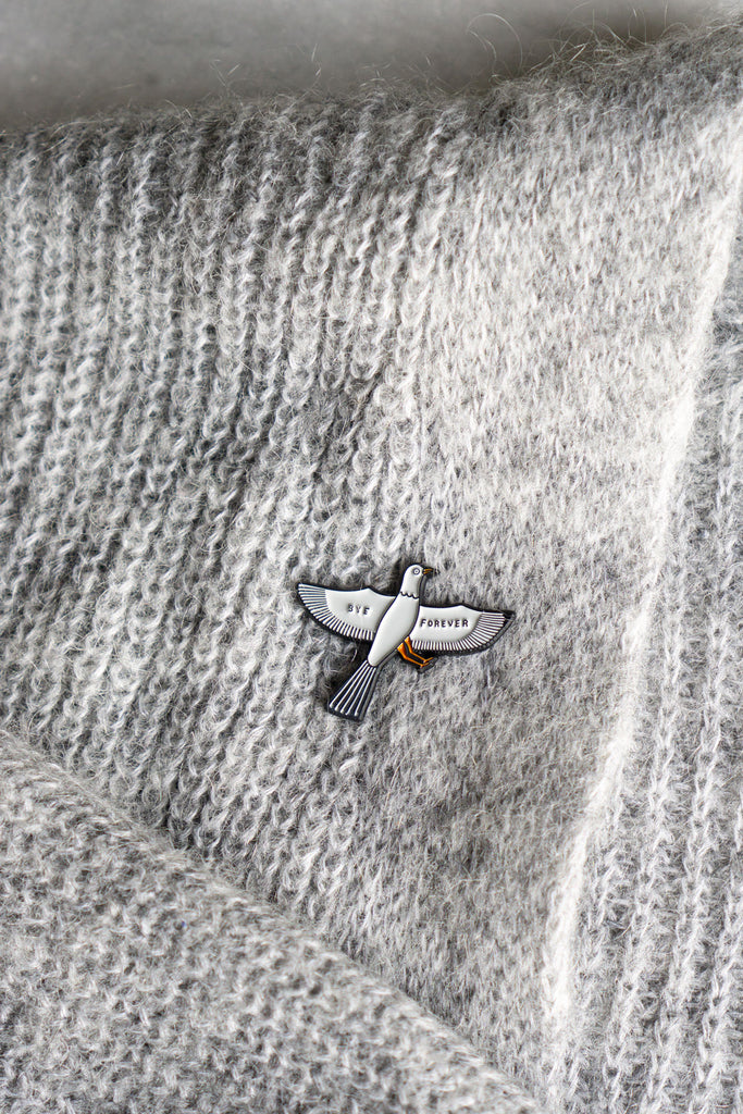 lapel pin of grey bird with wings outstretched and the text "Bye" and "Forever" on each wing pinned onto grey knitted sweater