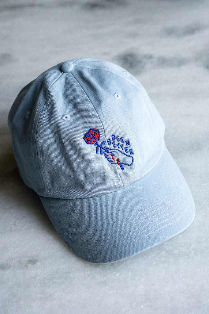 pale blue baseball hat with dark blue outline of hand with two red scratches holding red rose with blue text saying been better