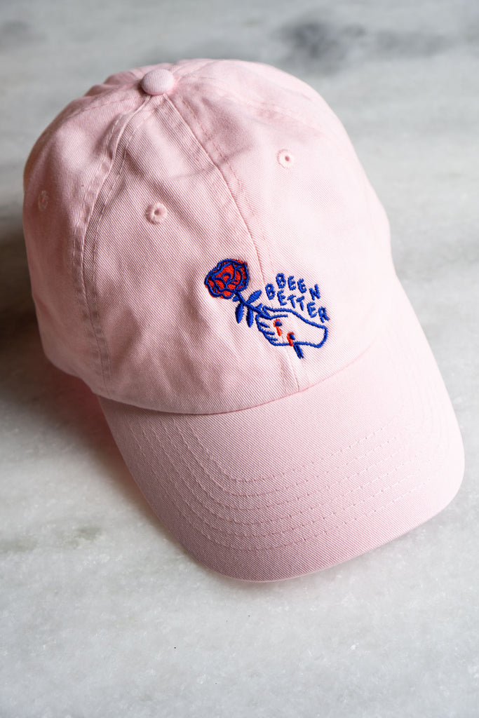 pale pink baseball hat with dark blue outline of hand with two red scratches holding red rose with blue text saying been better